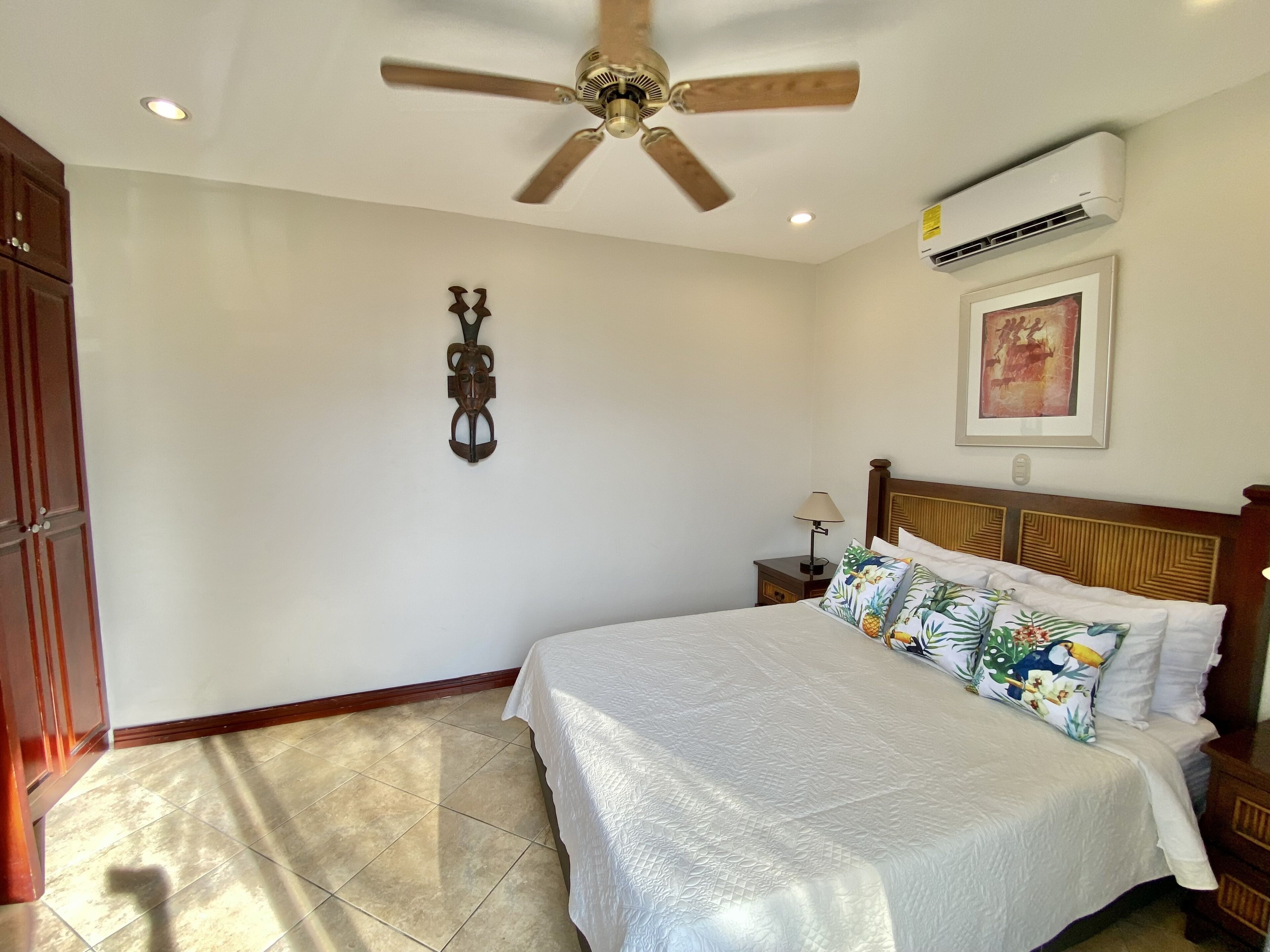 A bedroom with a bed, a ceiling fan, an air conditioning unit, a nightstand, and a wall-mounted decorative item.