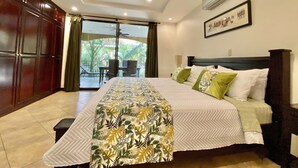 Un dormitorio con cama, ventilador de techo, aire acondicionado, ventana con vista a palmeras y puerta que da a un balcón.