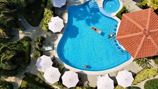 Una piscina grande y de forma peculiar con varias secciones, rodeada de sombrillas y sillones blancos.