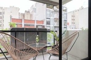 Un balcón con sillas de mimbre y una mesita pequeña, con vistas a un paisaje urbano con edificios y zonas verdes.