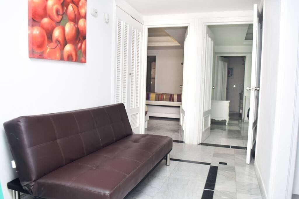 A modern living room with a brown sofa, a painting of red apples, and a hallway leading to a bedroom.