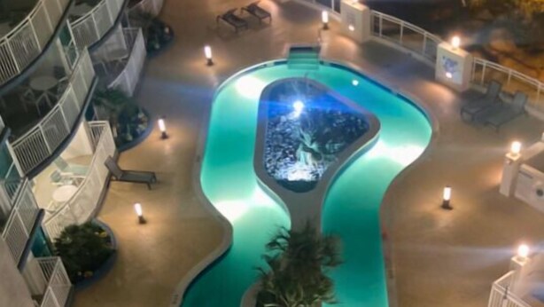 A pool with a unique wavy design, surrounded by lounge chairs and potted plants, with a building in the background.