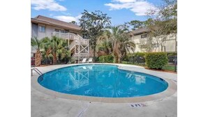 A swimming pool with a wooden deck, surrounded by palm trees and a building with a balcony.