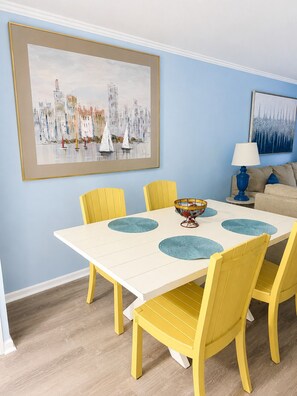 Un coin repas avec une table blanche, des chaises jaunes et un tableau encadré représentant des voiliers accroché au mur.