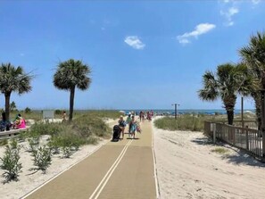 A sandy pathway with palm trees on both sides, leading to a beach area with people and beach umbrellas.