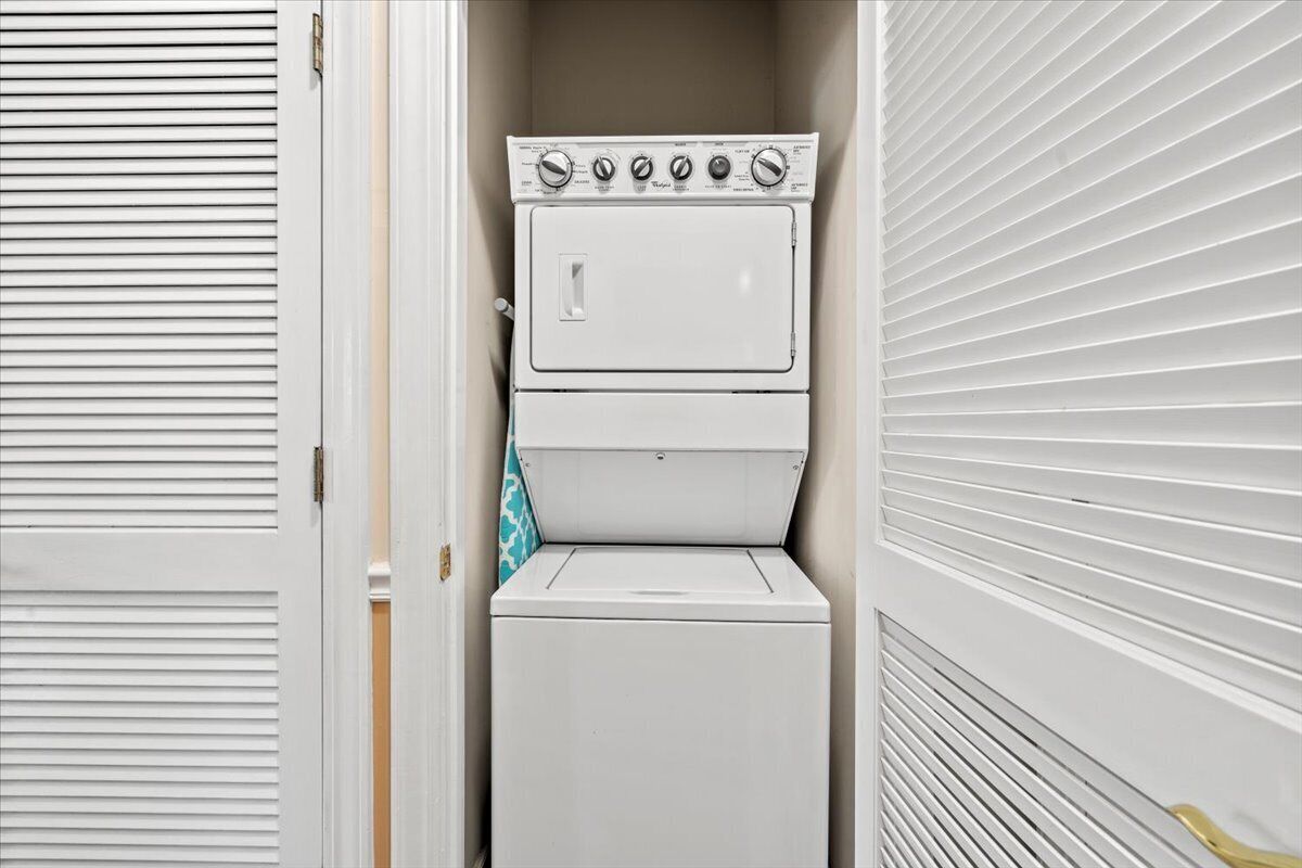 Een kleine wasruimte met een gestapelde wasmachine en droger, een handdoekhouder en een deur met horizontale zonwering.