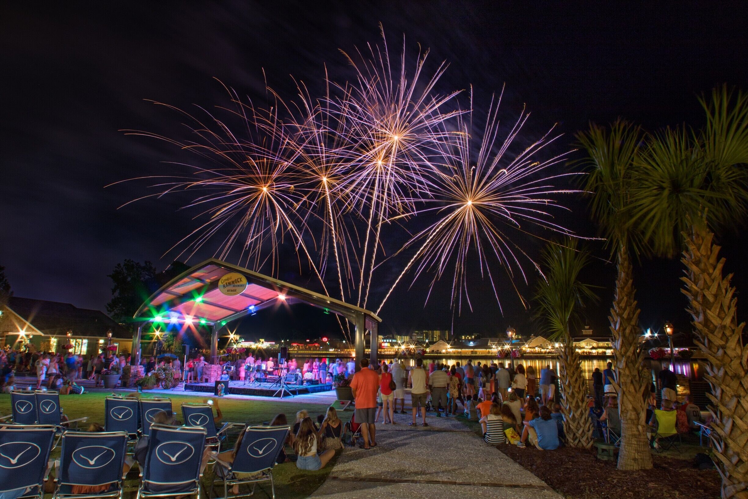 A covered outdoor seating area with rows of chairs, a stage with performers, and a view of fireworks in the background.