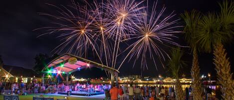 A covered outdoor seating area with rows of chairs, a stage with performers, and a view of fireworks in the background.