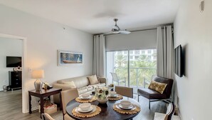 A modern living room with a dining table set for four, a sofa, a TV, and a large window with a view of palm trees. - Pet Friendly in Orlando area near Disney (Celebration)