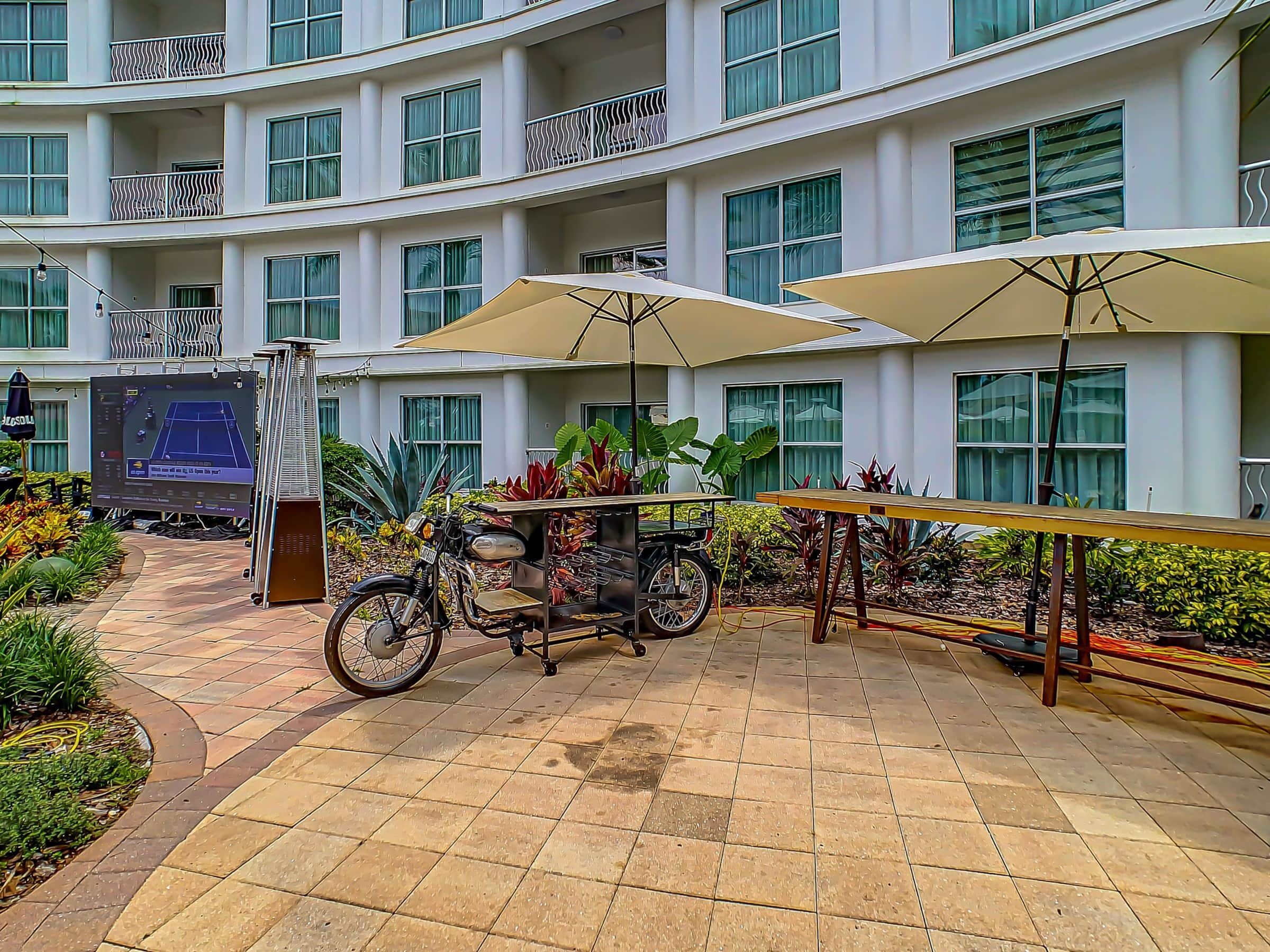 A courtyard with a tiled floor, a bicycle, a table with an umbrella, and a building with balconies and windows.