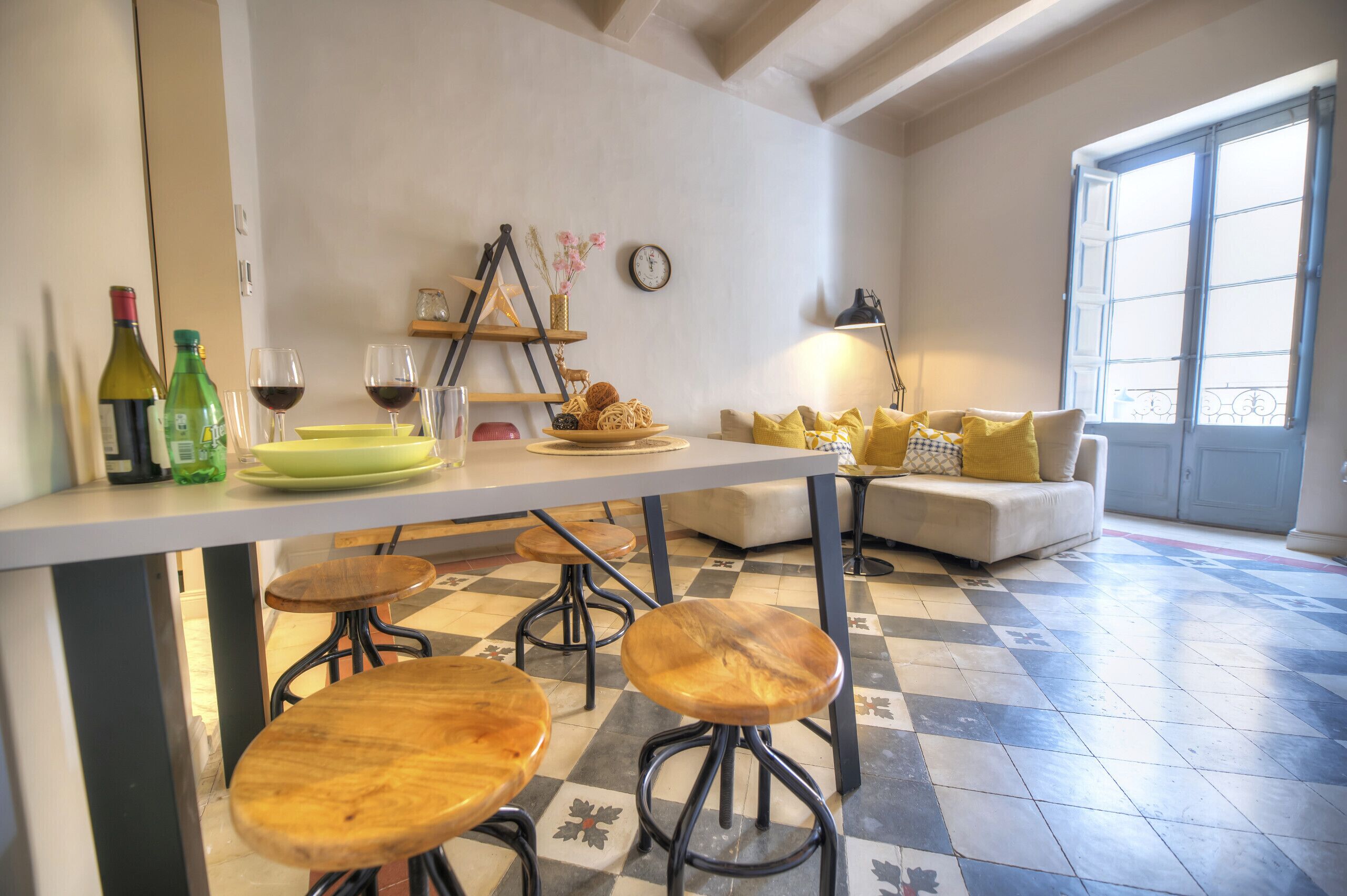 A kitchen with a dining table, stools, and a sofa with yellow cushions. There are wine bottles and glasses on the counter.