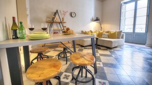 A kitchen with a dining table, stools, and a sofa with yellow cushions. There are wine bottles and glasses on the counter. - Charming & traditional apartment in Valletta ESCH2 (Valletta)