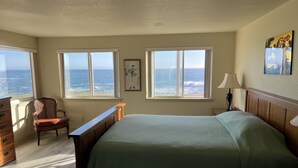 Une chambre à coucher comprenant un lit, une chaise, une commode et une table de chevet. Trois fenêtres offrent une vue sur l’océan.