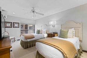 Quarto de hotel com duas camas, um ventilador de teto, uma mesa de cabeceira, um abajur e uma janela com vista.