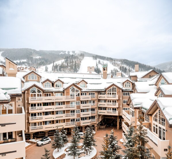 Hotel de vários andares com um pátio central, rodeado por árvores cobertas de neve e uma pista de esqui ao fundo.
