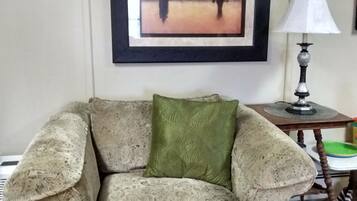 Una sala de estar con un sillón estampado, un cojín verde, un cuadro en la pared y una mesa con lámpara.