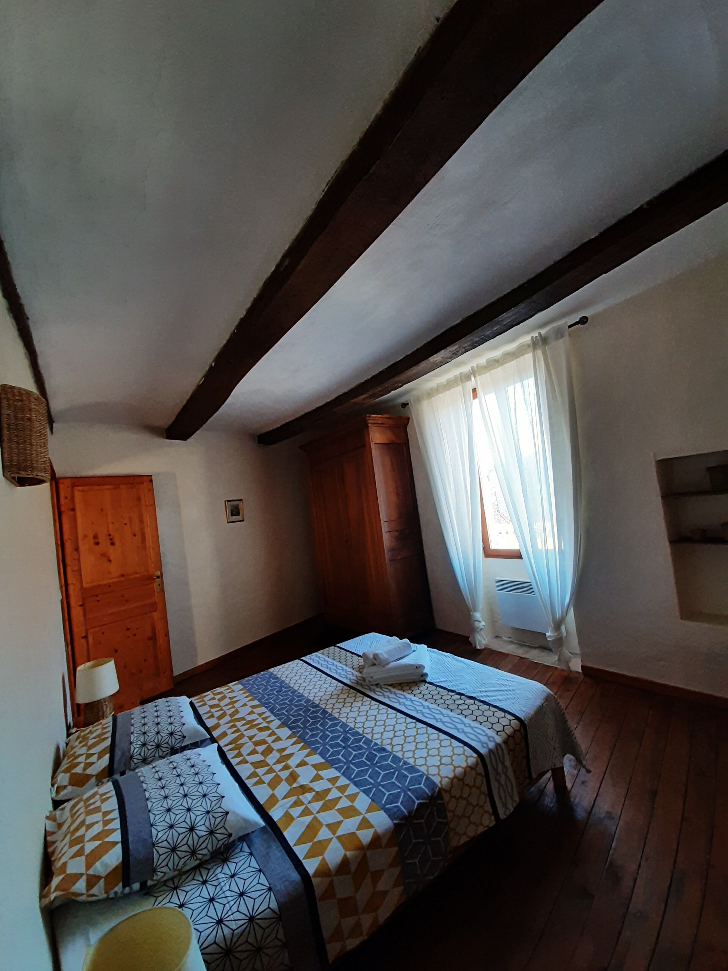A bedroom with a bed, wooden ceiling beams, a window with curtains, and a wall-mounted air conditioning unit.