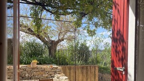 Une terrasse avec une table ronde en bois et des chaises, entourée de murs en pierre et d’un grand arbre.