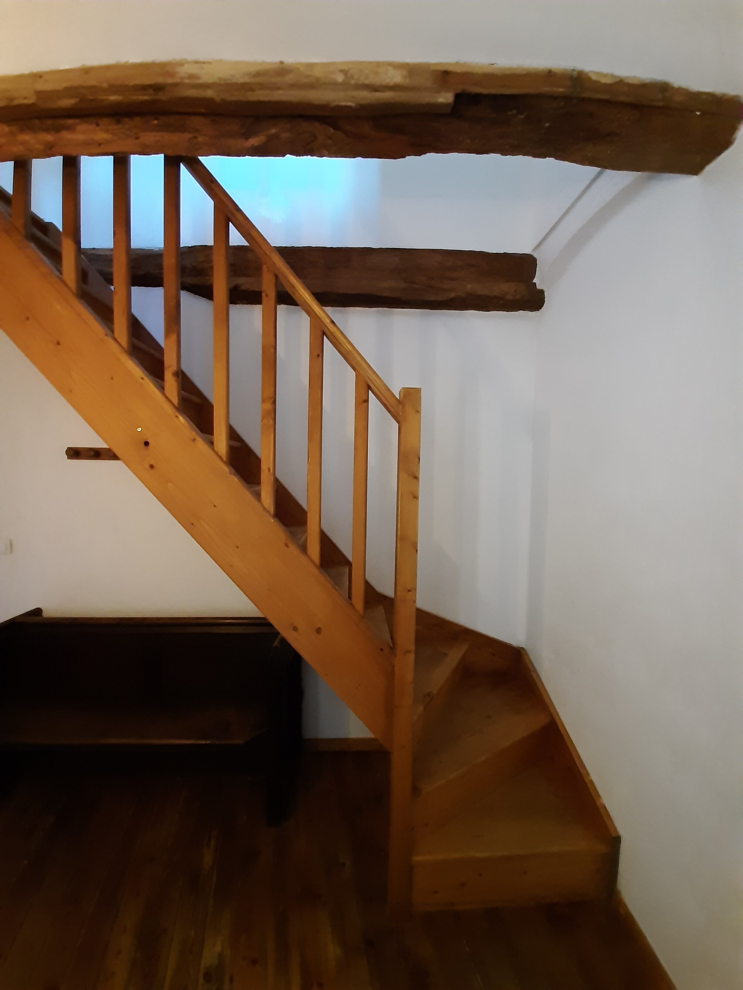 A wooden staircase with a rustic ceiling beam, a dark wooden bench, and a white wall.
