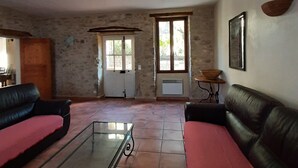 Un salon avec deux canapés en cuir noir, une table basse en verre, des murs en pierre, une porte en bois et une fenêtre donnant sur un paysage.