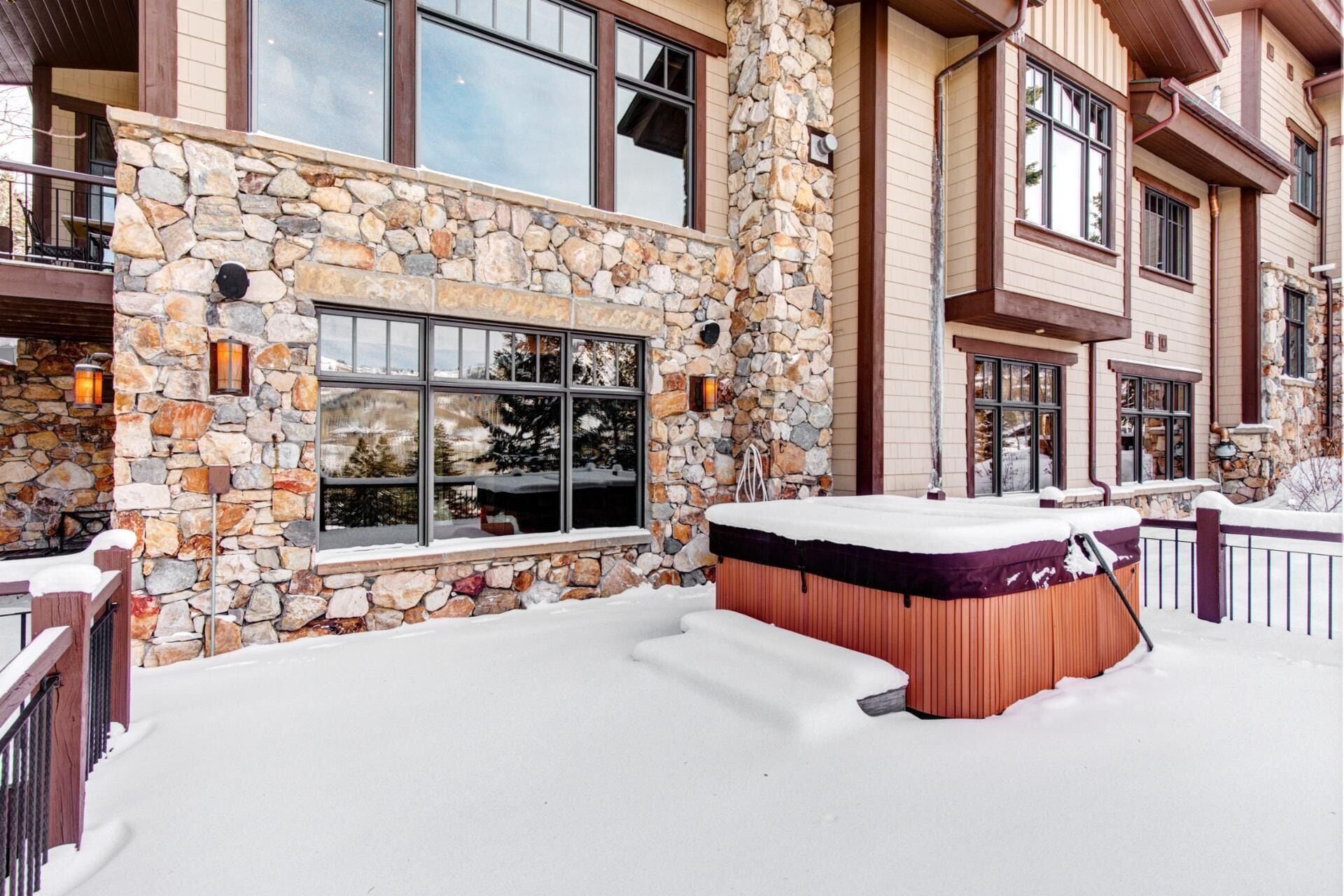 A stone exterior with large windows, a hot tub, and a snow-covered deck.