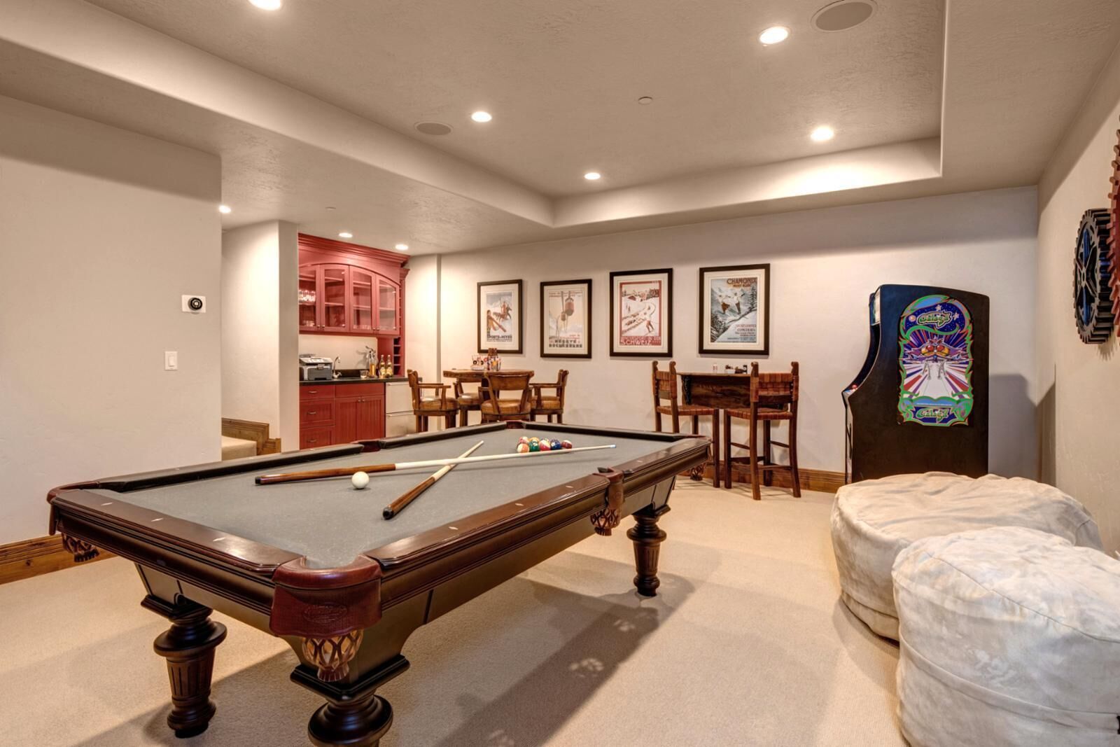 A pool table in a room with a kitchen area, framed pictures on the wall, and a dartboard.