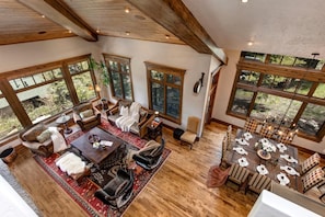 A spacious living area with a large wooden dining table set for six, surrounded by checkered chairs. There is a cozy seating area with a sofa and armchairs, and a fireplace. Large windows provide views of the surrounding landscape. - Lookout Drive 5 Bedrooms PLO-6599 by KBM (Park City)