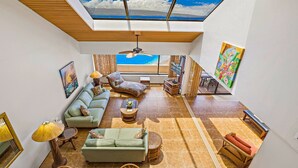 A spacious living area with a high ceiling, a ceiling fan, a large window with a view, and a painting on the wall.