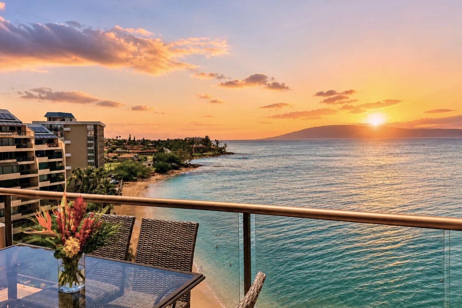A balcony with a view of the ocean at sunset, a table with chairs, and a tropical flower arrangement.