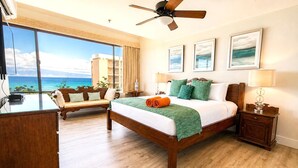 A hotel room with a bed, a desk, a chair, a ceiling fan, and a large window with a view of the ocean.