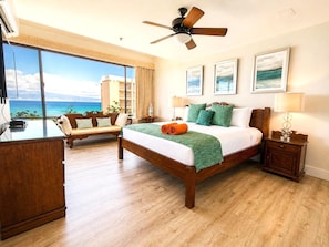 A hotel room with a bed, a desk, a chair, a ceiling fan, and a large window with a view of the ocean. - Sands of Kahana Resort by KBM (Lahaina)