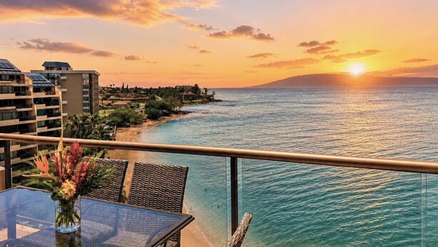 A seaside view from a balcony with a table and chairs, overlooking a beach and ocean.