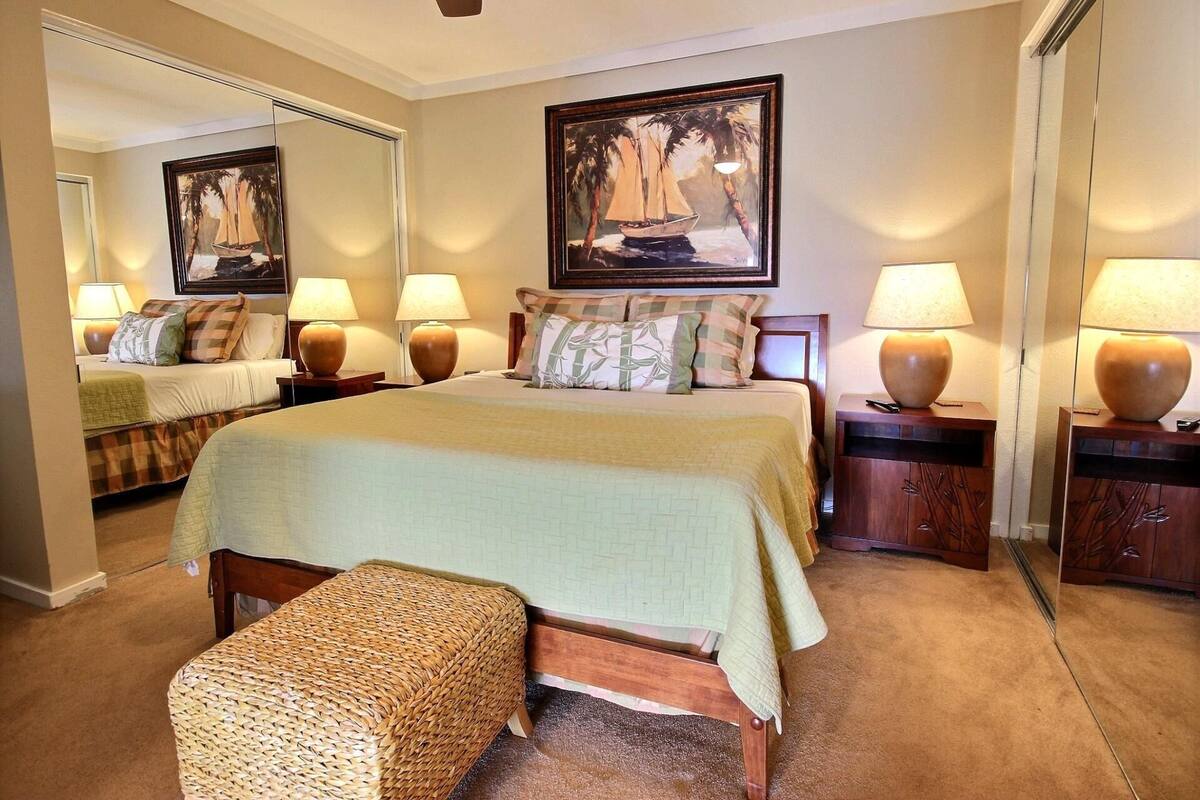 A well-lit bedroom with a bed, two bedside tables with lamps, a woven bench, and a painting on the wall.