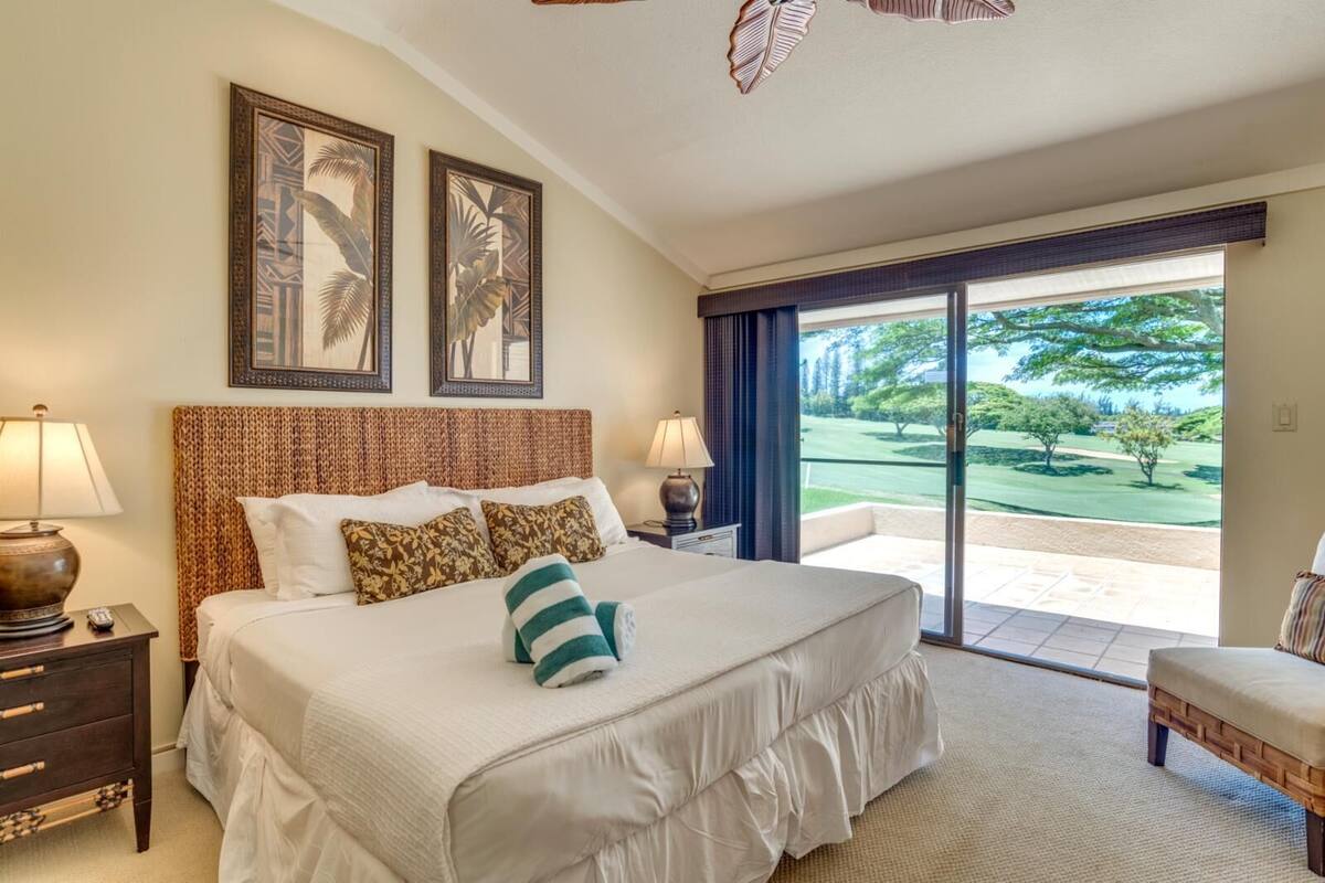 A hotel room with a large bed, two wall-mounted lamps, a nightstand, a wooden bench, and a view of a golf course through a sliding glass door.