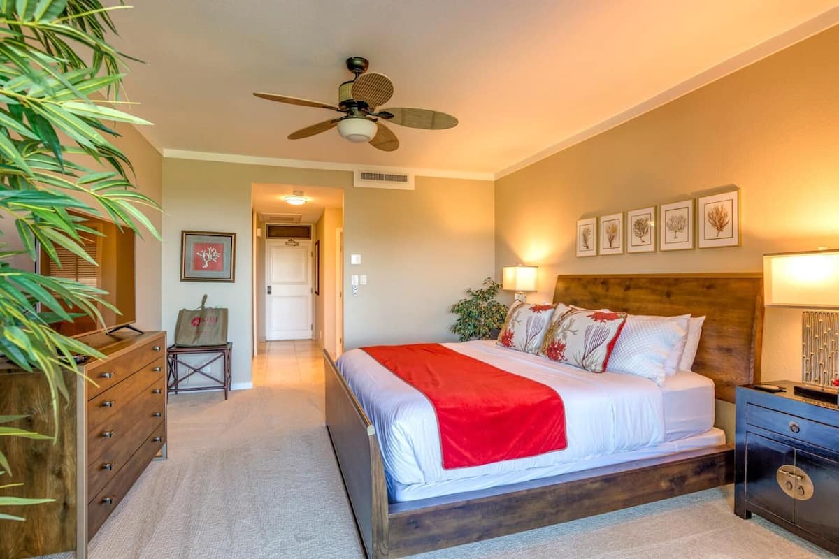 A bedroom with a bed, a wooden headboard, a nightstand, a dresser, a ceiling fan, and a door leading to another room.