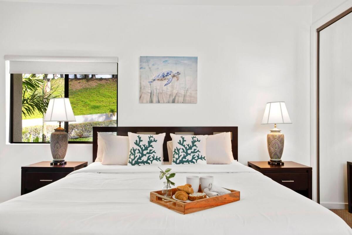 A neatly made bed with a tray of pastries, two bedside lamps, and a painting on the wall.