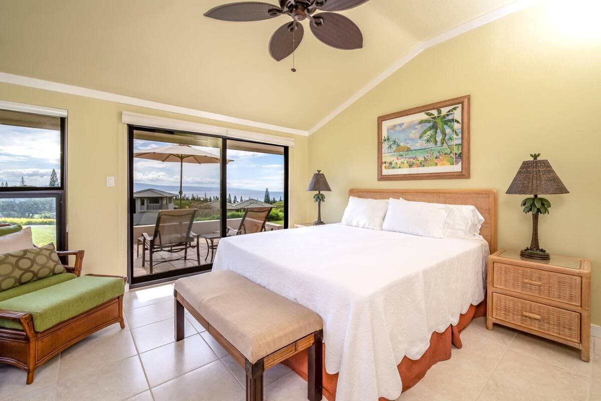 A bedroom with a large bed, a bench, a nightstand, a lamp, and a painting of a tropical scene.