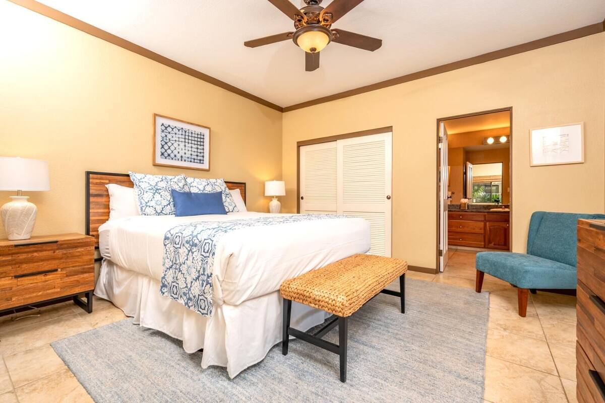 A bedroom with a bed, bedside tables, a bench, a ceiling fan, a door, and a chair.