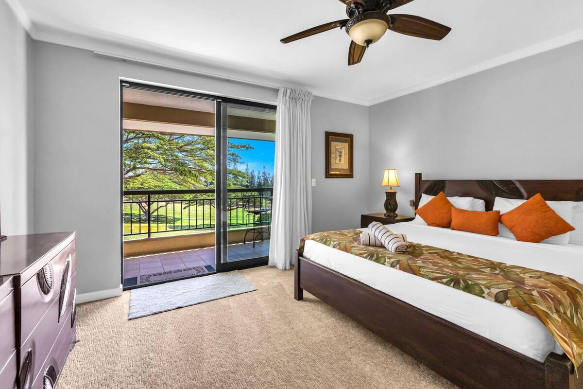 A hotel room with a bed, a nightstand, a ceiling fan, a window with a view of a green landscape, and a sliding door to the balcony.