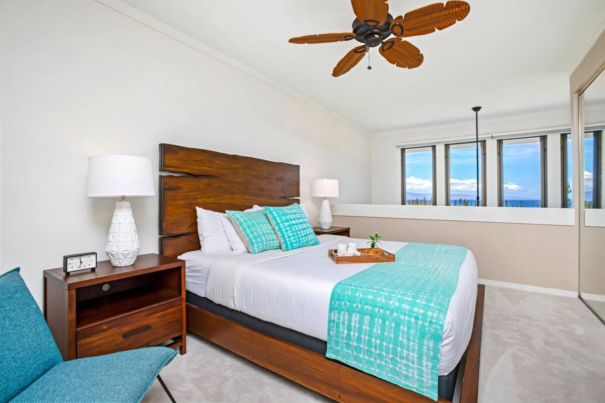 A bedroom with a wooden headboard, two bedside tables with lamps, a bed with a teal blanket, a nightstand with a clock, and a view of the sky through large windows.