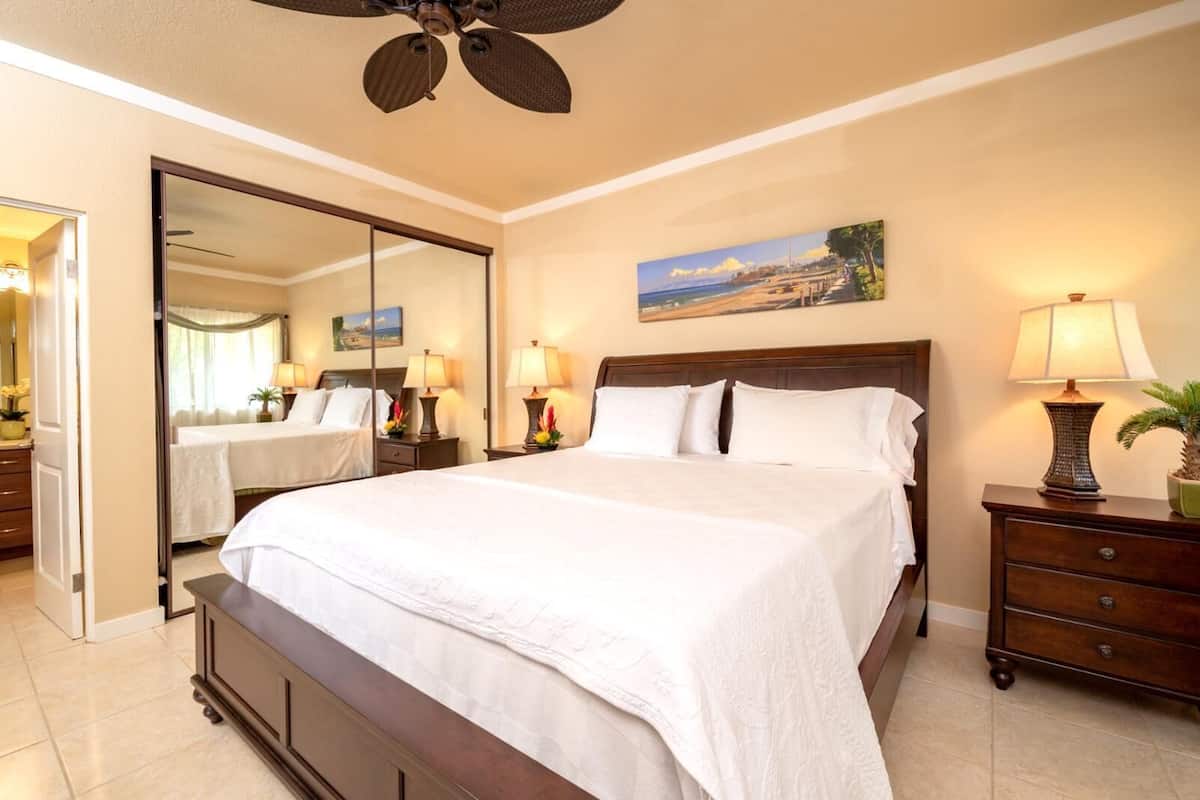 A hotel room with a large bed, a ceiling fan, a painting of a beach, and a nightstand with a lamp.
