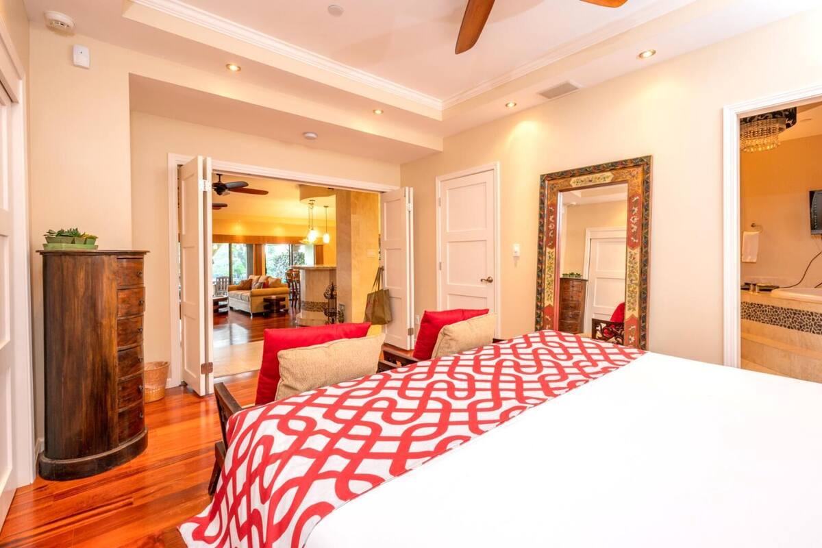A bedroom with a large bed, a wooden dresser, and a view into a living area with a sofa and dining table.