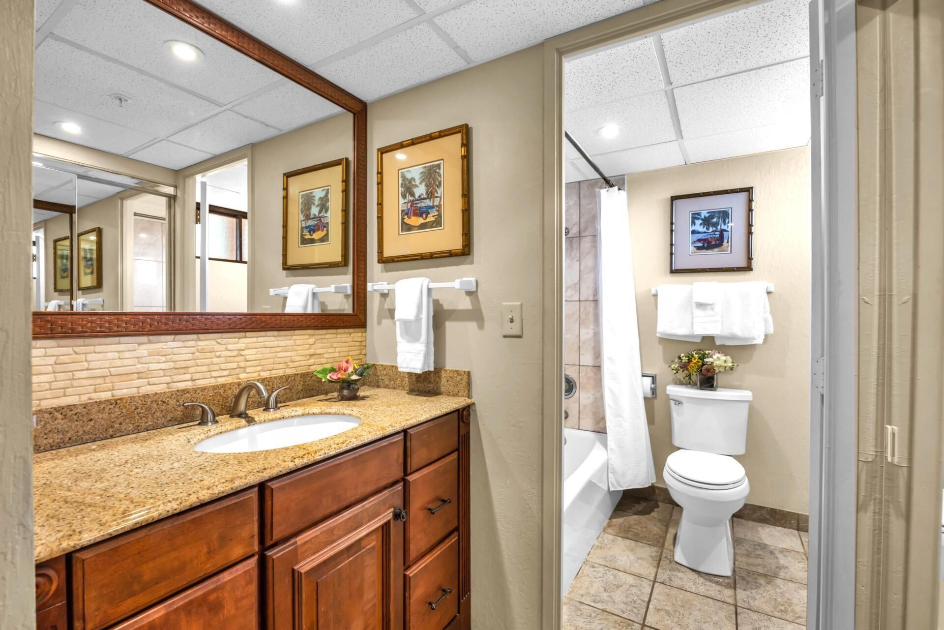 A bathroom with a granite countertop sink, wooden cabinets, a large mirror, framed pictures on the wall, a toilet, and a shower area.
