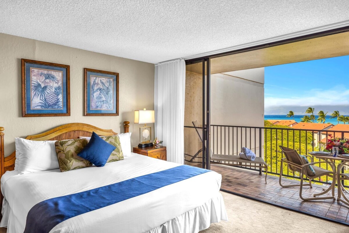 A hotel room with a bed, a nightstand, a lamp, two framed pictures, and a balcony with a view of the ocean and palm trees.