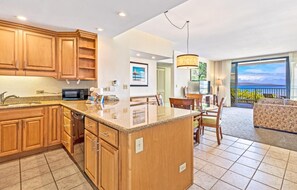 A modern kitchen with wooden cabinets and a central island, a dining area with a table and chairs, and a balcony with a view. - Kaanapali Shores Resort by KBM (Lahaina)