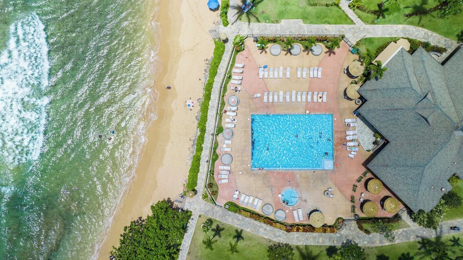 Ett flygbildsbild av ett semesterområde med en swimmingpool, en strand och ett område med solstolar och sittplatser.