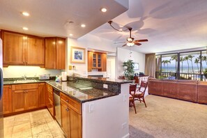 A modern kitchen with wooden cabinets, granite countertops, and a central island. There is a dining area with a view of the beach and palm trees. - Kaanapali Shores Resort by KBM (Lahaina)
