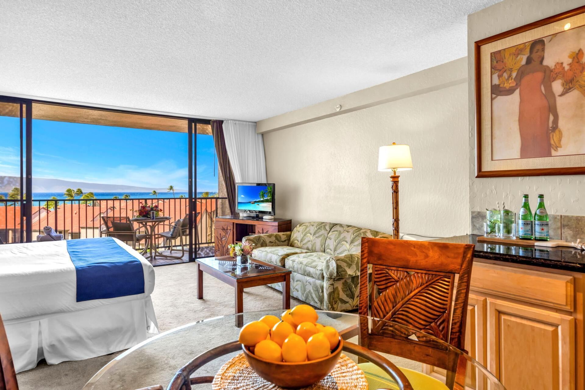 A hotel room with a bed, a dining table with a bowl of fruit, a TV, a sofa, and a balcony with a view.
