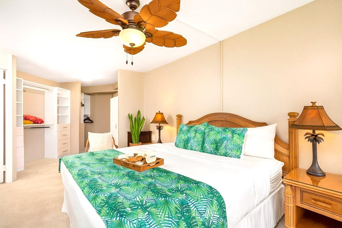 A bedroom with a bed, a ceiling fan, a nightstand, a lamp, a bedside table, a wardrobe, and a plant.