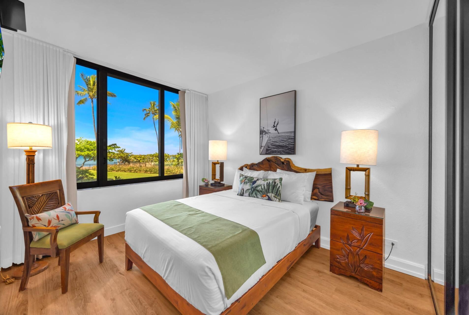 Une chambre d’hôtel avec une grande fenêtre, un lit recouvert d’un plaid vert, deux tables de chevet avec des lampes, une chaise agrémentée d’un coussin à motifs floraux et un tableau encadré accroché au mur.